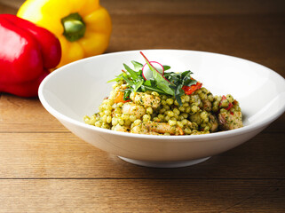 Homemade salad with pasta ptitim and vegetables, tomatoes, greens on wood background. Top view. Space for text. Red and yellow pepper in the background. Traditional Israeli pasta.