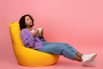 Millennial black female drinking coffee, relaxing in bean bag chair on pink studio background, copy space