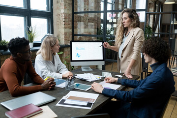 Pregnant businesswoman pointing at computer monitor with financial graphs and talking to her colleagues during business presentation at office