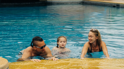 The mother and father with little daughter have fun in the pool. Mom and dad plays with the child. The family enjoy summer vacation in a swimming pool jumping, spinning, splash water. Slow motion.