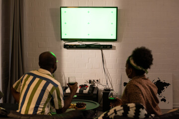 Rear view of African couple sitting on sofa with glasses of red wine and looking at widescreen TV...