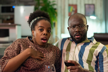 African young couple looking very frightened during watching horror movie with glasses of red wine, they sitting on sofa at home