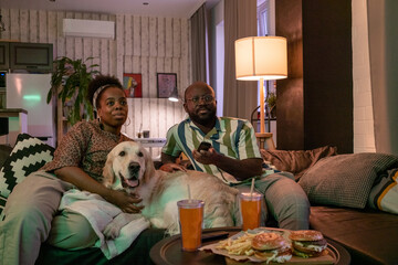 African young couple sitting on sofa with their dog, they watching TV and eating chips with burgers...