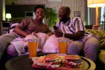 Close-up of burgers and soda on the table with African couple watching tv together on the sofa in the background