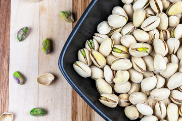 Black square bowl is filled with pistachios.  Bowl is angled on a butcher board background.  Additional pistachios scattered to the left.