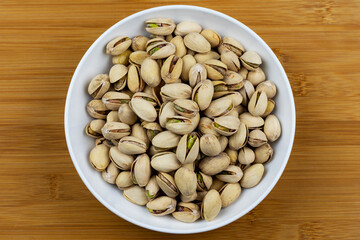 White bowl filled with pistachios centered on a bamboo background.