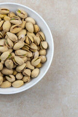 Top down view of a white bowl of pistachios on a tile surface.