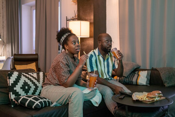 African young couple sitting on sofa drinking soda and eating fast food while watching the movie at home