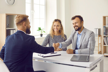 Happy young couple shaking hands with real estate agent after signing contract at his office. Clients smiling and exchanging handshake with realtor. Happy family thanking loan advisor for consultation - obrazy, fototapety, plakaty