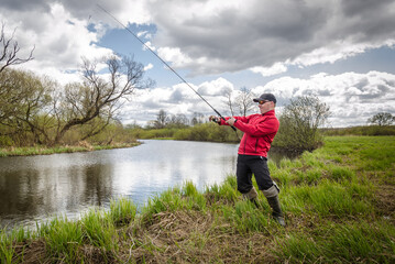 Fototapeta premium Fisherman in a red jacket is fishing on the green bank of the river.