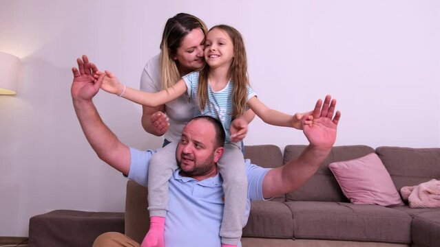 Happy family together spending time at home on weekend, girl sitting bihind on her father neck and making airplane position.