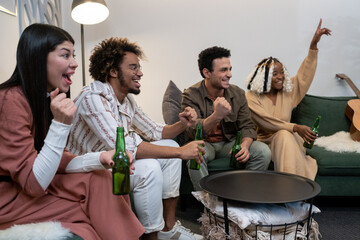 Excited young friends drinking beer and watching sport match while resting on sofa in the living room at home
