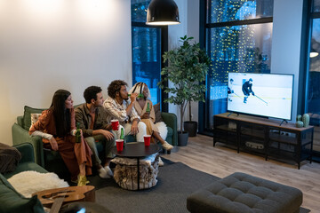 Naklejka premium Young friends sitting on sofa with beer and watching sport match on TV in a big modern apartment