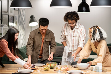 Group of young friends setting the dining table together putting the meal and plates, they preparing for dinner party