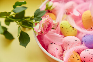 Fototapeta na wymiar Festive Easter background. Pink bowl with colorful eggs, pink feathers and pink rose. Happy Easter concept. Post card on yellow background