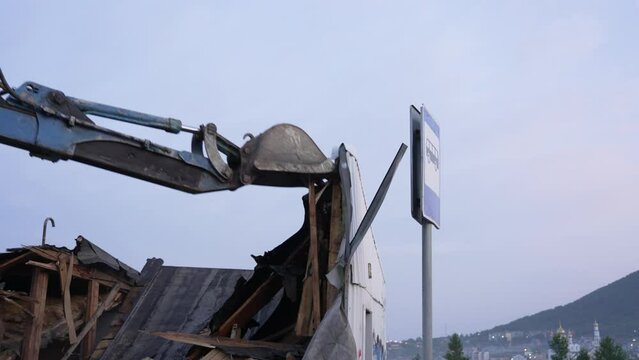 blue excavator using a bucket breaks a wooden abandoned house