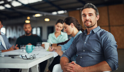 The creative process. Portrait of a mature man sitting at a table in an office with colleagues working in the background.