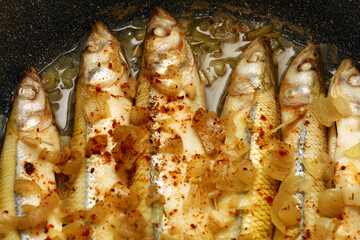 Delicious appetizing fresh fried european smelt fish with white onion and red seasoning on black non-stick frying pan, top view close-up. Six fish lie side by side, cooking at home