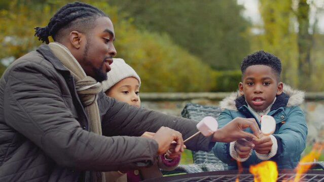 Father helping children as Family toast marshmallows sitting around firepit or barbeque in garden at home - shot in slow motion