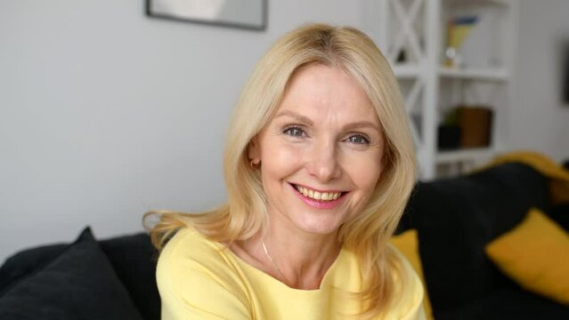 Senior woman in home interior. Charming mature caucasian lady looking at the camera and laughing, resting at the sofa. Serene female spends weekend at home