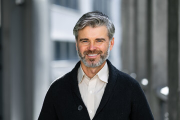 happy mature man looking at camera on the street. Senior businessman with a beard feels confident. Close-up face of a business man smiling on the background of the building.