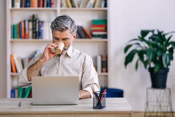 Senior man works on a laptop while sitting in a room and drinking coffee