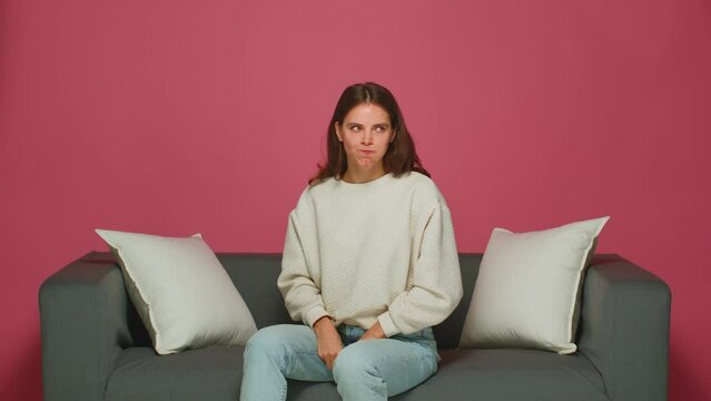 Funny playful young girl makes faces bulging ears showing tongue, teasing, fooling, sitting on sofa on pink background