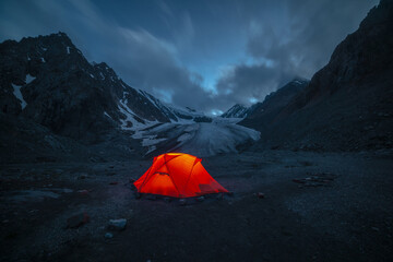 Awesome mountain landscape with vivid orange tent near large glacier tongue under clouds in night starry sky. Tent glow by orange light with view to glacier and mountains silhouettes in starry night. © Daniil