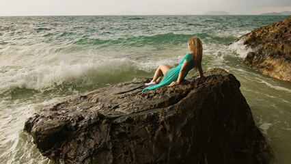 Seductive long-haired woman sitting on rock of sea reef stone, stormy cloudy ocean. Woman in blue...