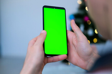 Closeup shot of man  hand using mobile phone with Green screen against the backdrop of a Christmas tree. Finger swiping up and down in green screen of the mobile. Green screen mockup template .