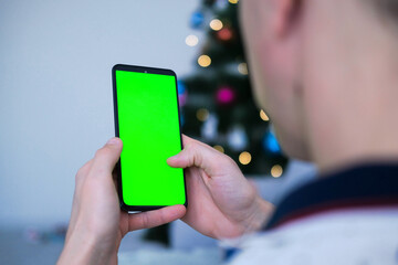 Closeup shot of man  hand using mobile phone with Green screen against the backdrop of a Christmas tree. Finger swiping up and down in green screen of the mobile. Green screen mockup template .