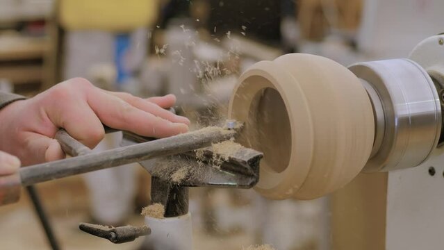 Close up: man carpenter using chisel for shaping piece of wood on turning lathe machine with many shavings, making wooden mug at workshop. Carpentry, hobby, craftsmanship and manufacturing concept