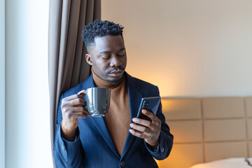 Businessman standing by window in hotel room drinking coffee. Thoughtful young African businessman standing by window with a cup of coffee and looking at his mobile phone.