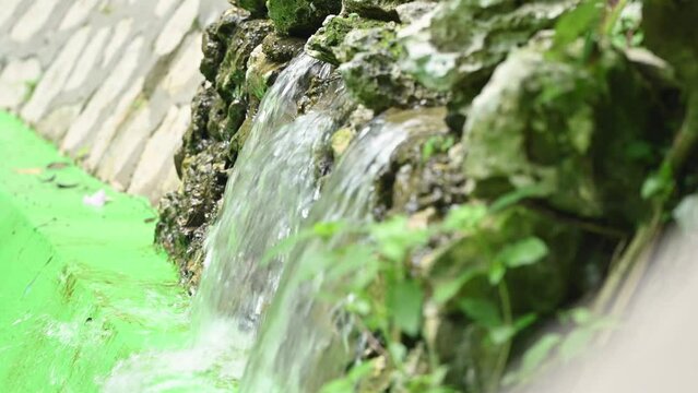 Fountain and flowing water closeup. Flowing water between river stones in natural lagoon. Small pond with fountain. Source of crystal water and drops reflect sun's rays. Pure aqua in movement. Park an