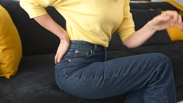 Cropped view of the senior woman felt a sharp pain in loin sitting on the sofa at home, worried caucasian lady holding a hand on the lower back