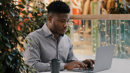 Focused young african american male freelancer carefully looking at laptop screen working remotely typing business email writing article chatting on social network online using computer application