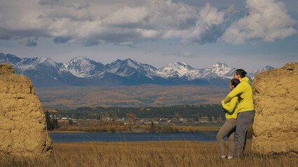 Man and woman in yellow green sportswear. Lovely couple of travelers hug and kiss near old stone enjoying highland landscape. Two travelers are walking against the backdrop of snow-capped mountains.