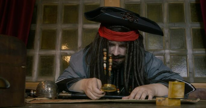 Pirate captain with dark long hair in cocked hat looks through magnifying glass on old sea map AT table searching for treasures in cabin