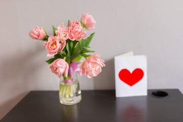 Bouquet of pink tulips in a glass vase with a postcard with a red heart. Romantic spring background