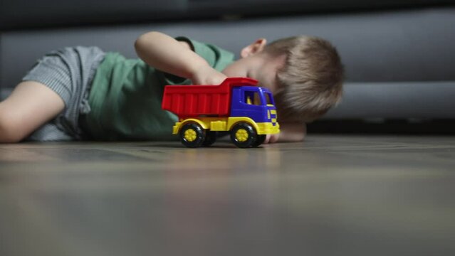 caucasian preschooler boy kid lying wooden floor in living room driving playing with plastic colorful toy car truck at home. 3 year child in green shirt doing creative activities pretending imagine 