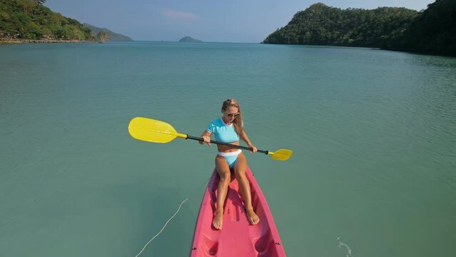 Pretty sportive blonde lady in stylish swimsuit sails on pink plastic canoe along azure ocean bay at exotic resort. Traveling to tropical countries. Girl is sailing on kayak in ocean, view from above.