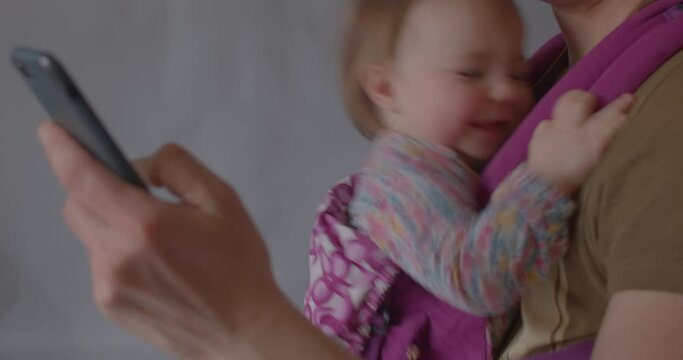 Cute little girl smiles coquettishly while sitting in an ergonomic baby carrier while her father surfs the internet, use phone. Periodic short-term use of sling and effect on hip development.