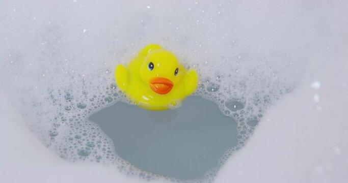Bright yellow rubber toy duck falls into warm water of bathtub with thick layer of bubbles and foam at bathroom illumination closeup