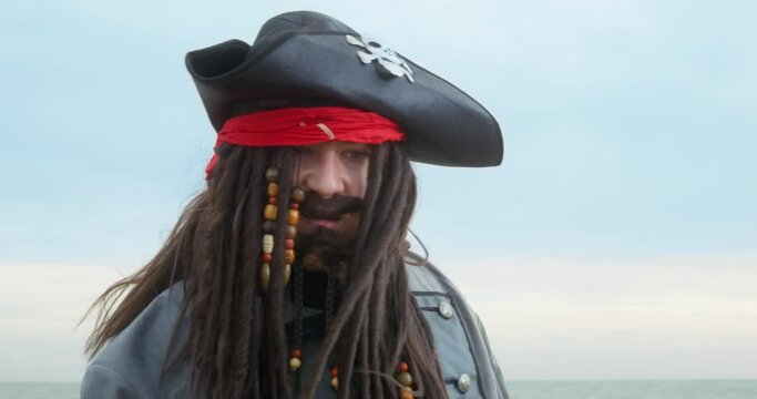 Sloppy angry pirate with cocked hat and dark long braids waves hands gesticulating on beach against sea IN cloudy weather closeup