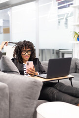 Brown curly young woman working happy with a phone and laptop on a sofa