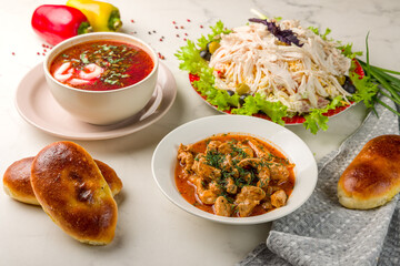 Goulash with beef and carrots, borscht with sour cream, chicken salad and pies on white marble table