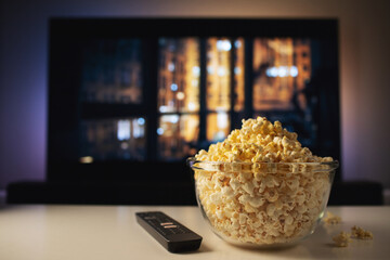 A glass bowl of popcorn and remote control in the background the TV works. Evening cozy watching a movie or TV series at home - obrazy, fototapety, plakaty