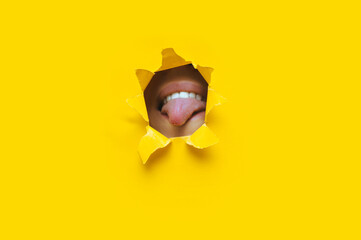 A young girl shows her tongue and teeth with lips. Torn hole in yellow paper. Copy space. The...