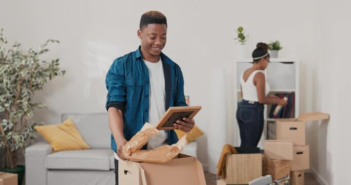 Handsome man unpacks cardboard boxes with things from the move, takes out a frame with a picture from the old days, a childhood photo, shows his wife, they dissolve in memories, laughter, joy