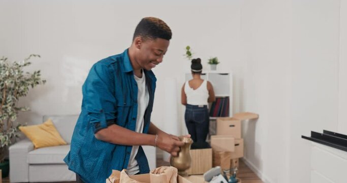 A handsome man is unpacking cartons of belongings from the move, taking out packed items, in the background a beautiful woman is also cleaning, arranging books on shelves